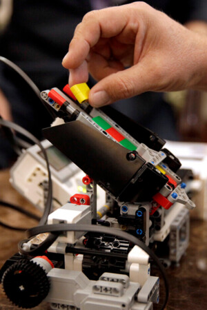 Uma mão a colocar uma peça num protótipo de um robot, pousado numa mesa, atrás vê-se a parte de baixo do tronco de uma pessoa, com uma mão na perna.